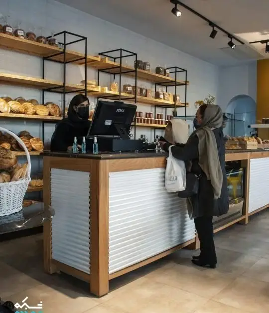 مشتری و کارکنان در کافه نانوایی پاتیس؛ زنی ماسک‌زده پشت پیشخوان به مشتری ماسک‌زده سفارش نان و شیرینی می‌دهد، قفسه‌های چوبی پر از محصولات.
