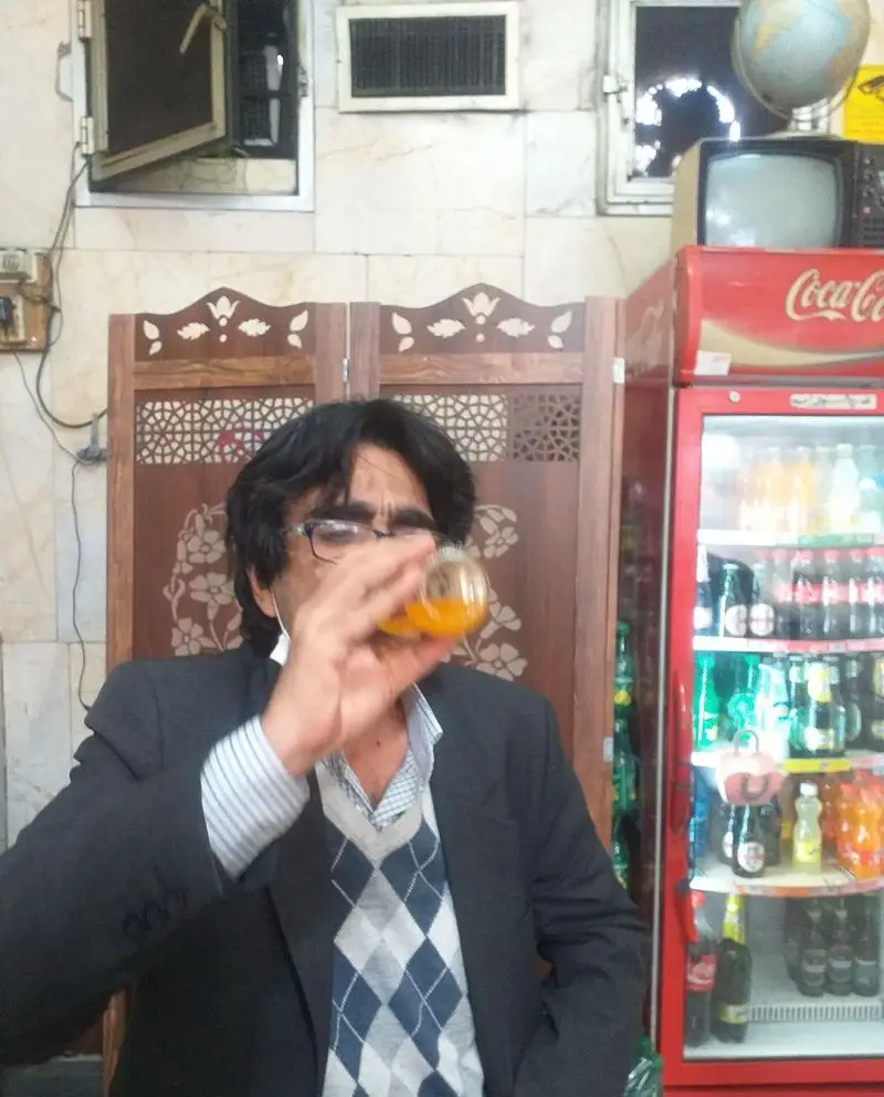 مردی در ساندویچ آذر در حال نوشیدن، در پس زمینه یخچال نوشابه و دکور چوبی دیده می شود که فضایی دلنشین ایجاد کرده است.