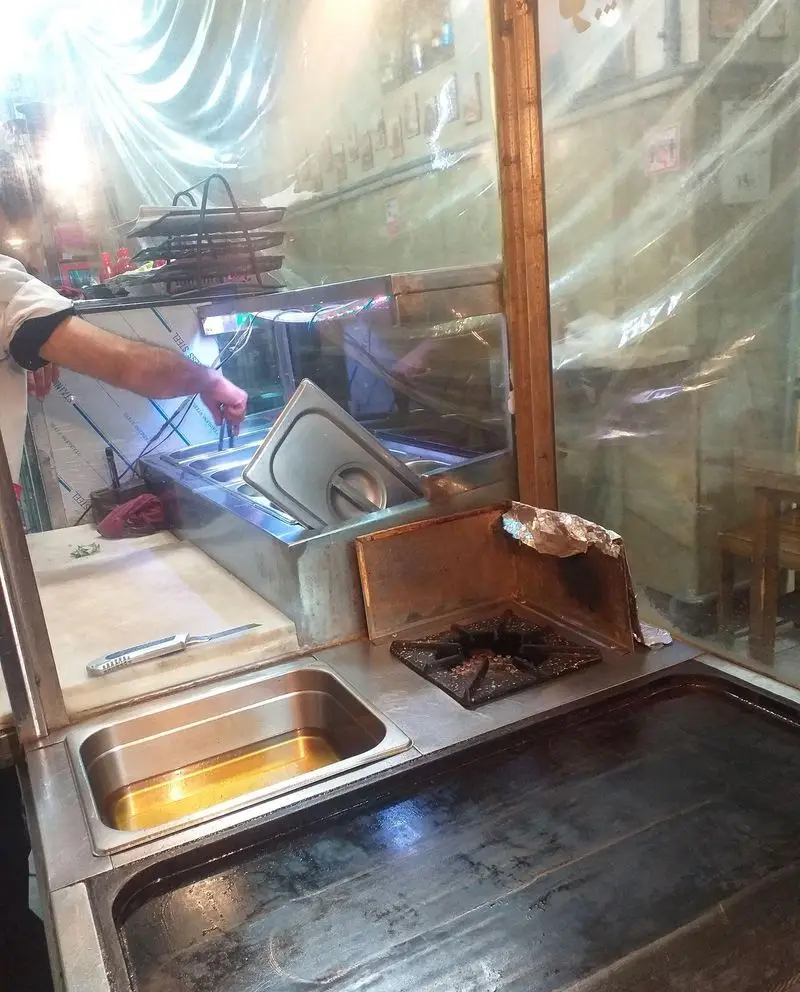 آشپزخانه ساندویچ آذر: آشپز در حال آماده‌سازی غذا در محیطی تمیز با تجهیزات پخت و پز مدرن شامل گریل و ظروف استیل برای ساندویچ‌های تازه و خوشمزه.