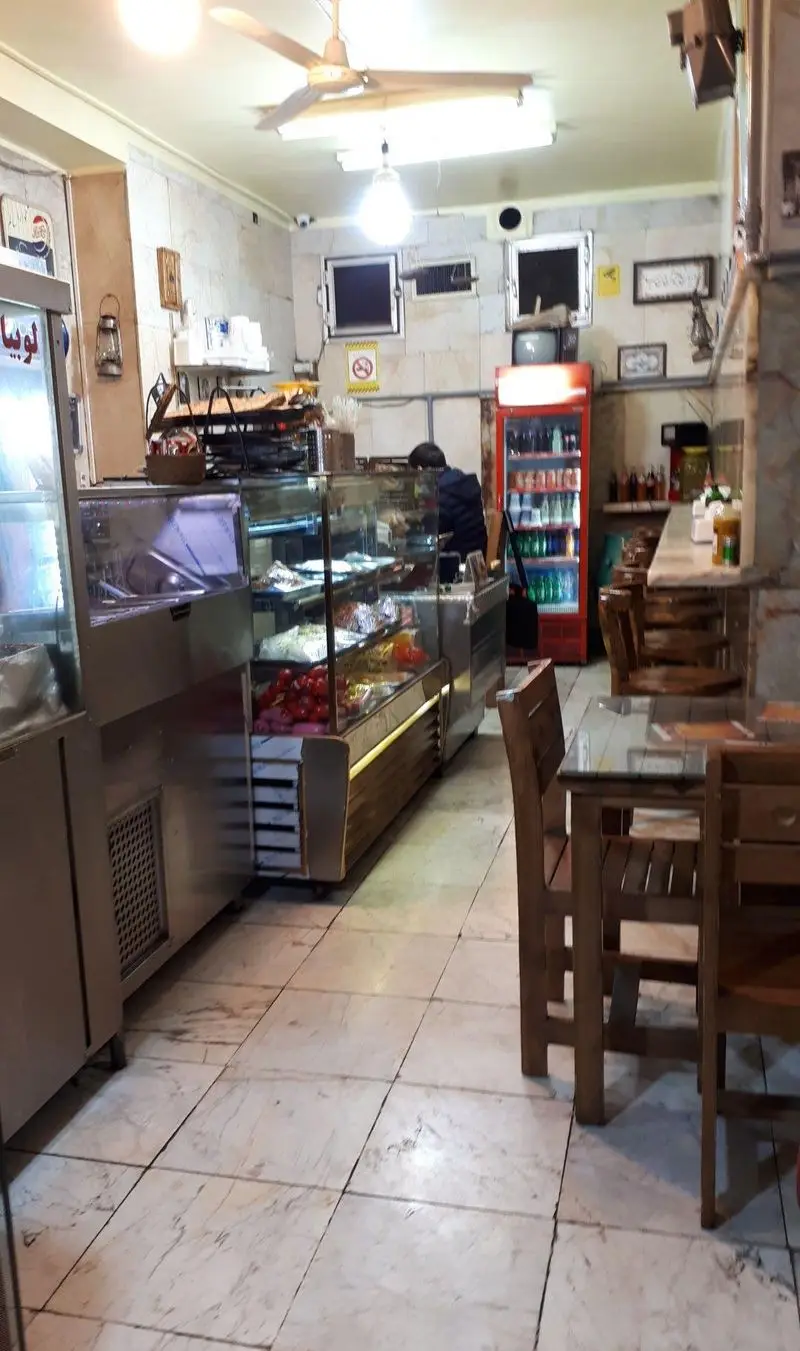 فضای داخلی ساندویچ آذر با ویترین‌های پر از ساندویچ و سالاد، یخچال نوشیدنی، میز و صندلی چوبی برای مشتریان. محیطی تمیز و آماده سرویس‌دهی.