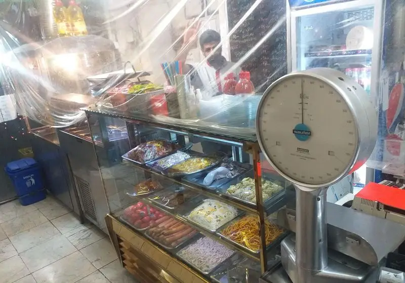 محیط داخلی ساندویچ فروشی آذر، با پیشخوان نمایش غذاهای آماده متنوع، شامل سالاد، سوسیس و پاستا. ترازوی Sauter و کارمند.