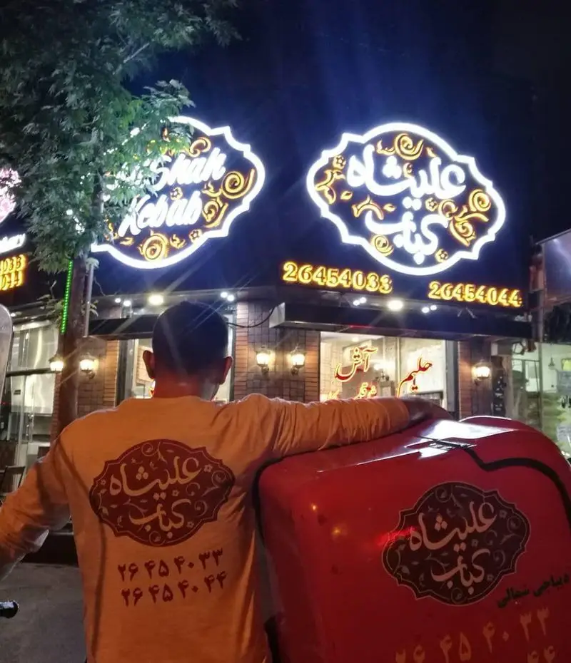 نمای شبانه رستوران کباب عالیشاه با تابلوهای نئون درخشان و شماره تماس ۲۶۴۵۴۰۳۳ و ۲۶۴۵۴۰۴۴. پیک عالیشاه با لباس فرم و باکس دلیوری قرمز رنگ آماده ارسال سفارشات.