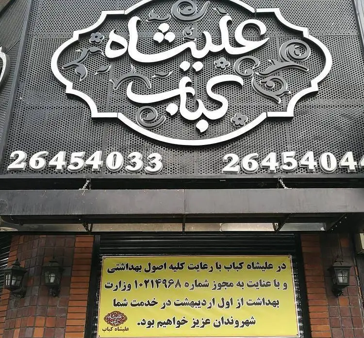 نمای بیرونی کباب عالیشاه، با تابلوی بزرگ، شماره‌های تماس 26454033 و 26454044 و بنر اطلاعیه بهداشتی وزارت بهداشت.