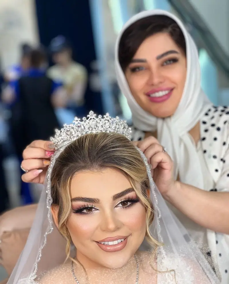 عکس شینیون موی عروس با تاج نقرهای پر زرق و برق و تور سفید در حال آمادهسازی نهایی توسط آرایشگر در سالن زیبایی مجلل