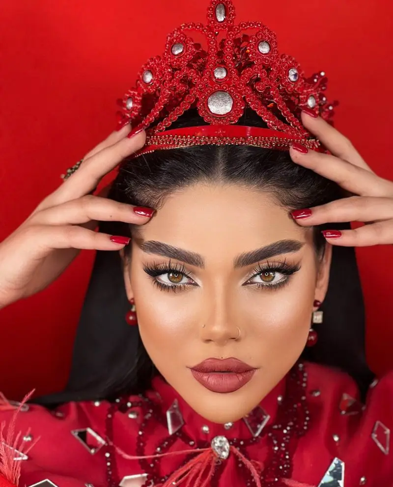 مدل زن با میکاپ کامل و غلیظ چهره، تاج مجلل قرمز روی سر و لاک قرمز ناخنها در سالن زیبایی نیلوفرآبی با پسزمینه سرخ