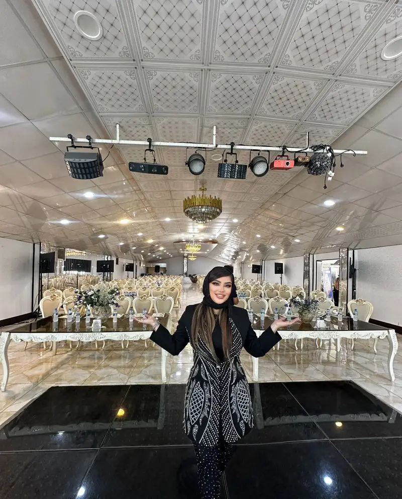 زن میکاپ آرتیست سالن زیبایی نیلوفرآبی در سالن بزرگ پذیرایی با صندلیهای سفید و تجهیزات نورپردازی سقف ایستاده است.