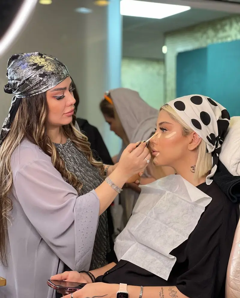 میکاپ آرتیست سالن زیبایی نیلوفرآبی در حال اعمال کرم پودر و کانسیلر روی صورت مشتری با روسری خالخالی سفید و مشکی