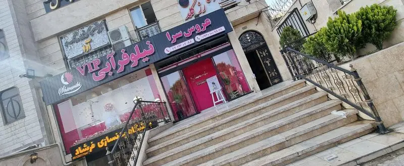 نمای بیرونی و ورودی ساختمان عروس سرای نیلوفرآبی با تابلوی سردر مشکی و صورتی، پلههای سنگی و شیشههای بزرگ ورودی سالن