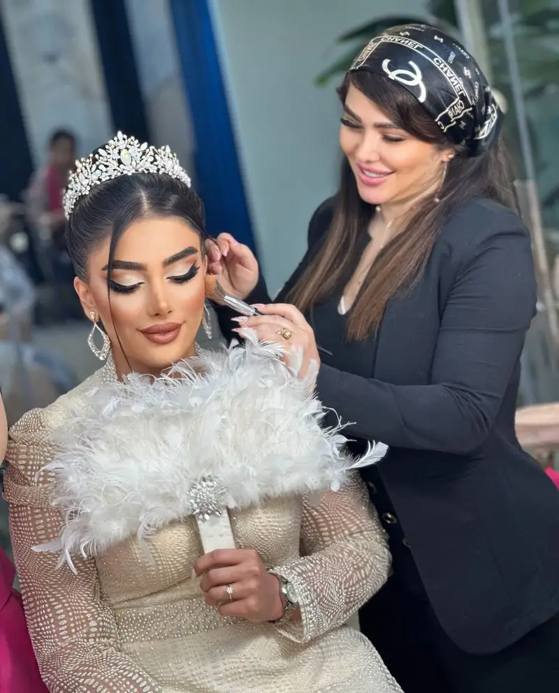 میکاپ آرتیست حرفهای در حال اعمال رژگونه روی صورت عروس زیبا با تاج درخشان، میکاپ لایت و بادبزن پر سفید در سالن زیبایی
