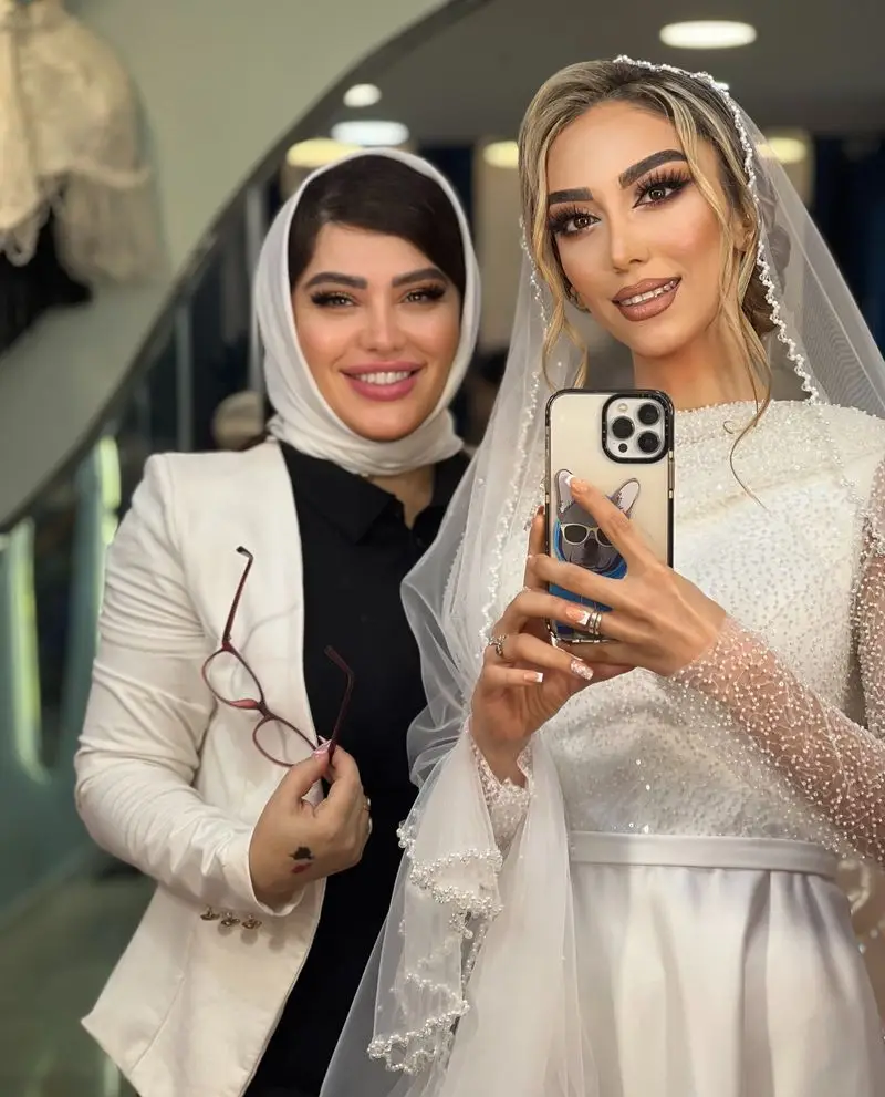 عروس با لباس تور سفید در حال گرفتن سلفی همراه با میکاپ آرتیست سالن زیبایی نیلوفرآبی با کت سفید و روسری مشکی.