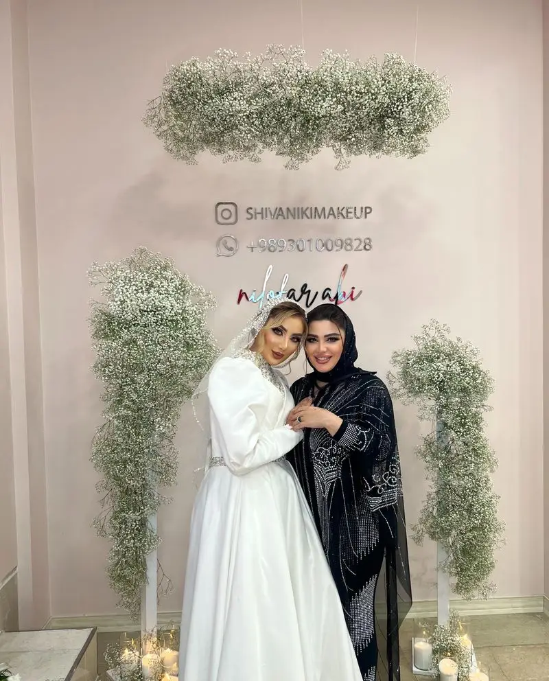 عروس با لباس سفید و میکاپ آرتیست با لباس مشکی در دکور گلآرایی شده با گلهای سفید ژیپسوفیلا در سالن زیبایی نیلوفرآبی