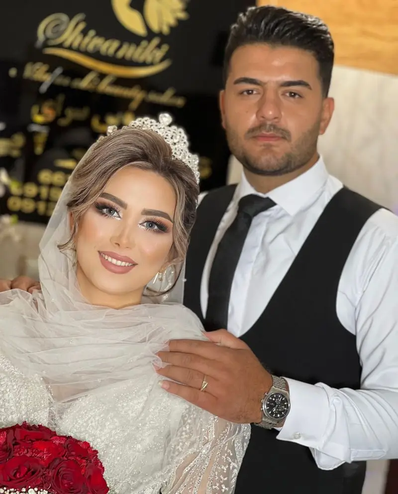 تصویر عروس با تاج درخشان و آرایش کامل چشم و لب همراه داماد با جلیقه مشکی و دسته گل رز قرمز در سالن زیبایی نیلوفرآبی.