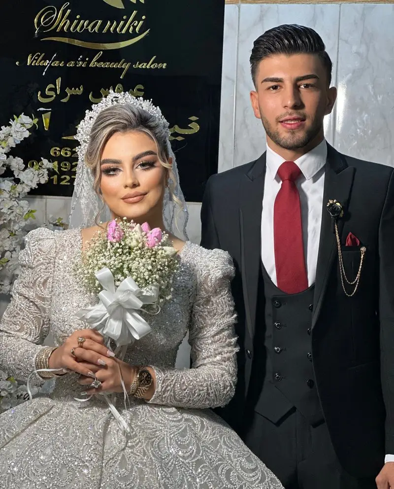 عروس و داماد در کنار سالن زیبایی نیلوفرآبی، عروس با لباس عروس پر زرق و برق و تاج زیبا، داماد با کت و شلوار مشکی و کراوات قرمز.