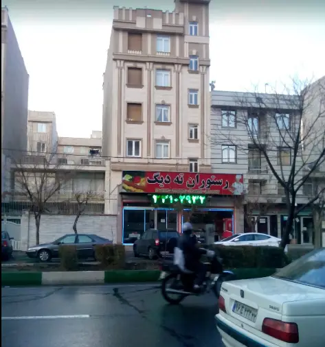تصویری از نمای بیرونی رستوران ته دیگ در یک خیابان شلوغ شهری. تابلو قرمز رستوران با شماره تماس سبز رنگ آن مشخص است. ماشین‌ها و موتورسیکلت در حال حرکت دیده می‌شوند.
