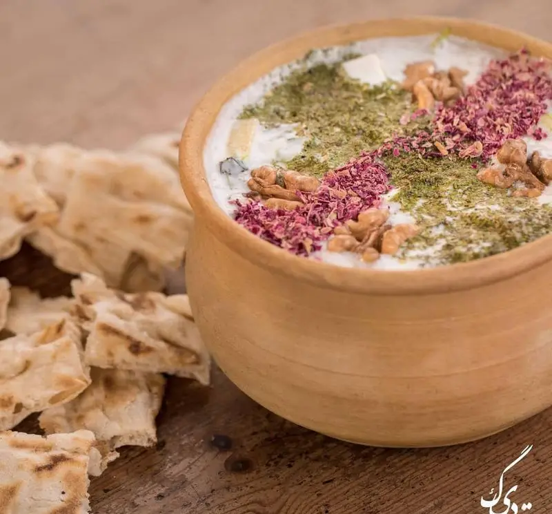 ماست و خیار سنتی رستوران ته دیگ در کاسه سفالی، تزئین شده با سبزی معطر، گردو و گل محمدی، همراه با نان تازه.