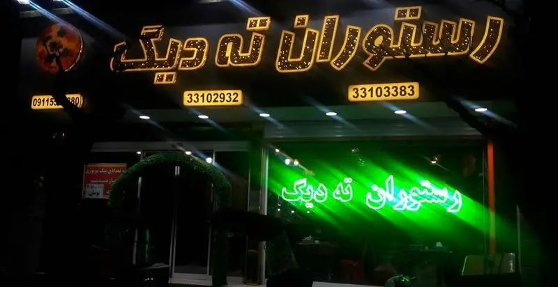 نمای بیرونی رستوران ته دیگ در شب با تابلوی درخشان، نئون سبز و شماره تلفن‌ها: 33102932، 33103383. تجربه غذای خوشمزه ته دیگ.
