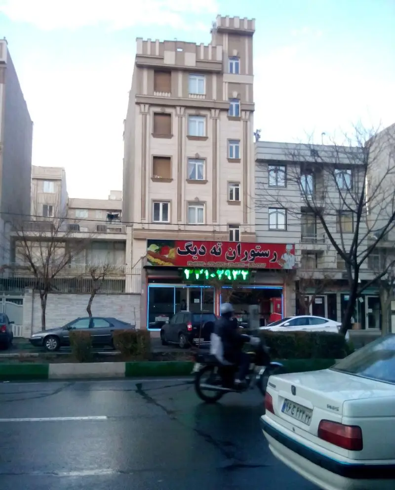 نمای بیرونی رستوران ته دیگ در یک خیابان شلوغ شهری با ماشین و موتور سیکلت. شماره تماس: ۲۲۱۰-۲۹۳۲ برای سفارش غذای ایرانی.