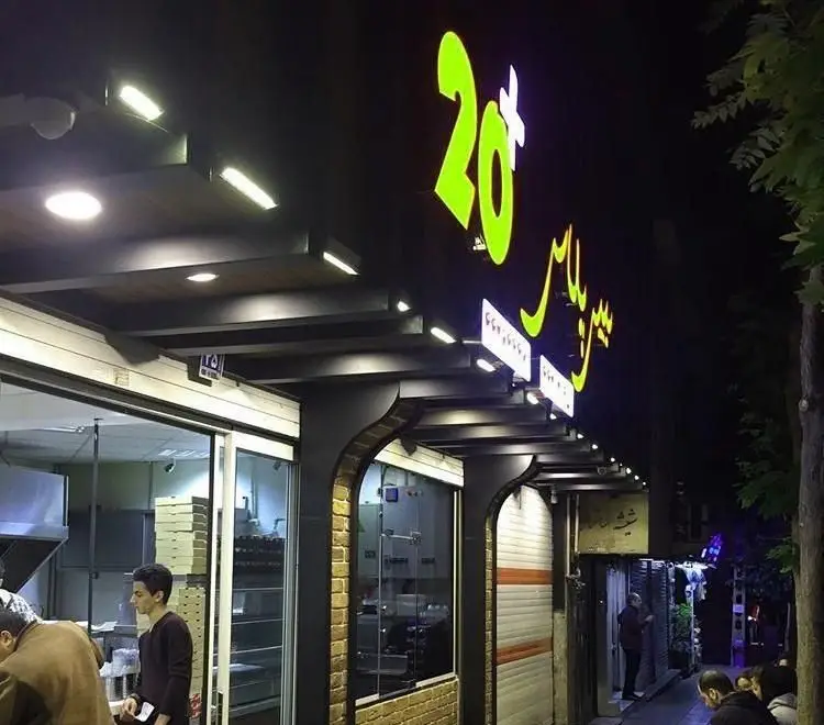 تصویری جذاب از نمای شبانه فست فود بیست پلاس، با نورپردازی روشن تابلوی 20+ و سیرپلاس، و حضور پر جنب و جوش مشتریان و پرسنل در ورودی.