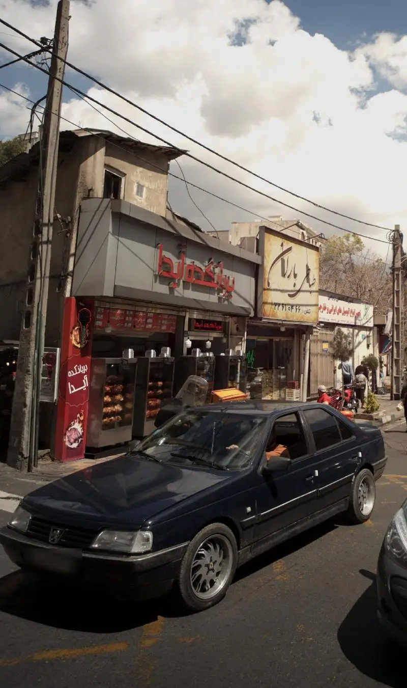 بریانکده آیدا با نمای مدرن خاکستری و قرمز، عرضه کننده مرغ بریان و غذاهای ایرانی، در خیابانی با سیم‌های برق و یک خودروی تیره.