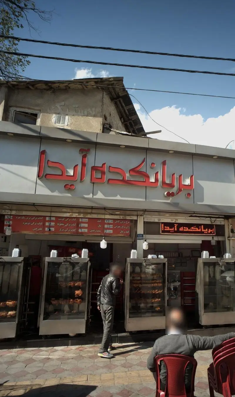 نمای بیرونی بریانکده آیدا، فروشگاه مرغ بریان و انواع کباب‌های لذیذ با تابلو قرمز رنگ، دستگاه‌های جوجه گردان و مشتریان در حال انتظار.