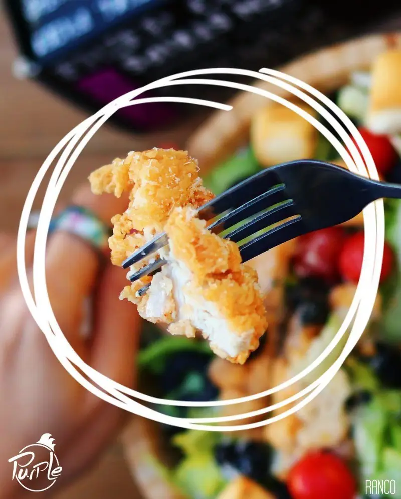 تکه مرغ سوخاری ترد و طلایی روی چنگال در حال جدا شدن، در کنار سالاد تازه پرپل سوخاری با سبزیجات، گوجه و زیتون