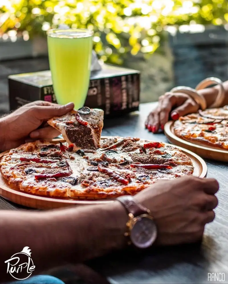 دست‌ها در حال برداشتن تکه‌ای از پیتزای خوشمزه و داغ پرپل سوخاری، در کنار نوشیدنی تازه، برای تجربه‌ای دلپذیر.