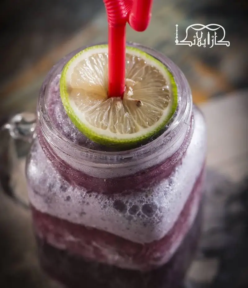نوشیدنی اسموتی بنفش خنک و خوشمزه با برش لیمو و نی قرمز در ماگ شیشه‌ای. تجربه طعم‌های خاص در کافه عربی کازابلانکا.