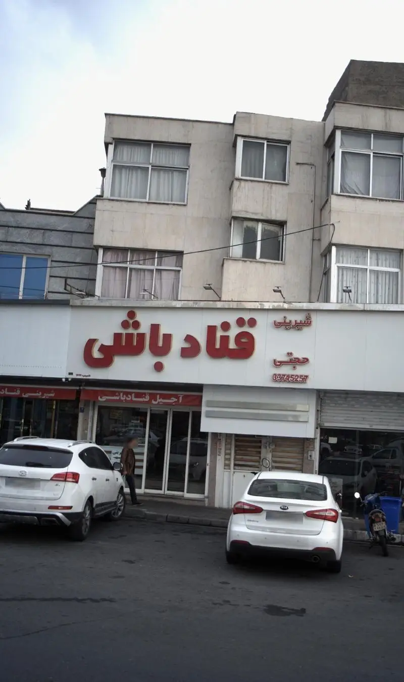 نمای بیرونی مغازه شیرینی و آجیل قناد باشی حجتی با تابلوی قرمز و واضح. دو خودرو سفید پارک شده در مقابل فروشگاه.