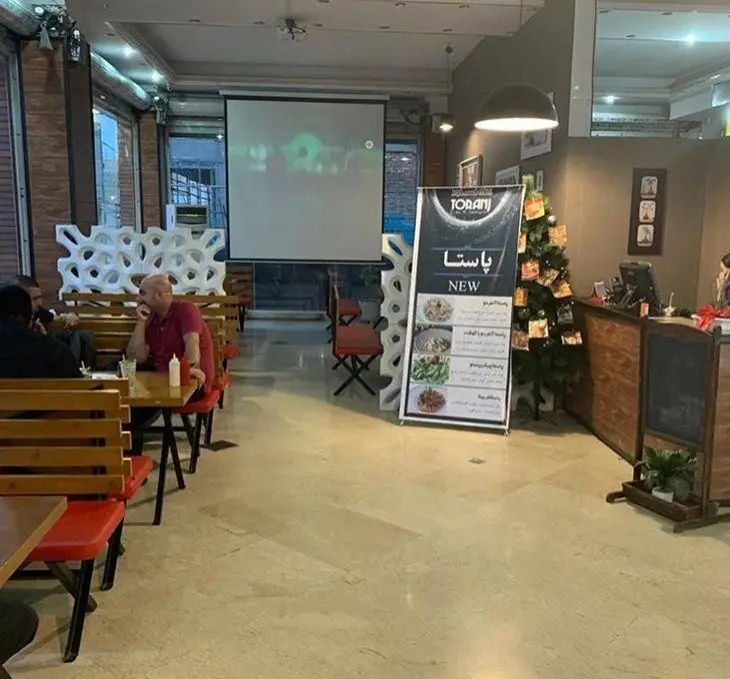 فضای داخلی فست فود ترنج شعبه ۳، با مشتریان، پروژکتور، استند تبلیغ پاستا و دکور کریسمس، محیطی دنج و مدرن.