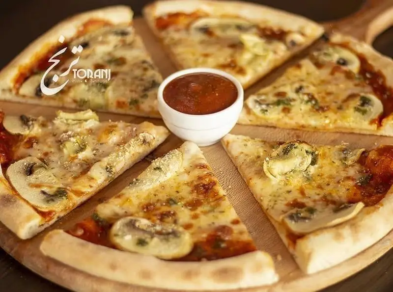 پیتزای قارچ و پنیر تازه و خوشمزه از فست فود ترنج شعبه ۳، برش خورده روی تخته چوبی چیده شده، همراه با سس مخصوص در مرکز.