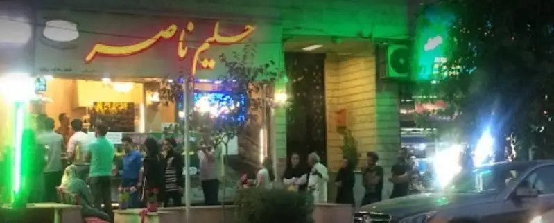 نمای شبانه از آش و حلیم پزی حلیم ناصر، با تابلوی نئونی درخشان و صف مشتریان مشتاق برای آش و حلیم تازه و باکیفیت.