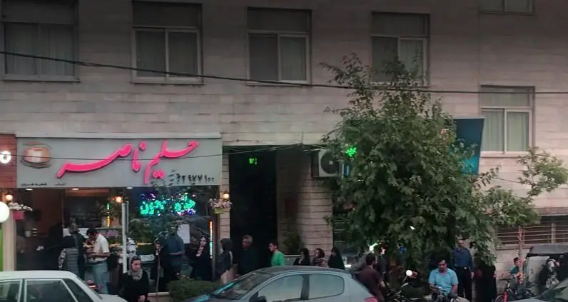 تصویر نمای بیرونی شلوغ آش و حلیم پزی حلیم ناصر در تهران، با مشتریان و وسایل نقلیه در حال خرید حلیم و آش داغ.