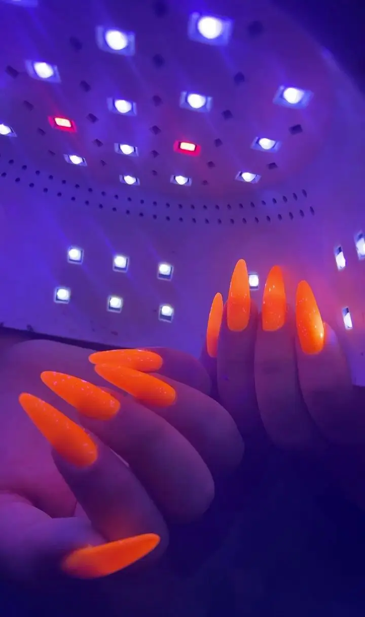 طراحی و کاشت ناخن بلند نوک تیز با لاک ژل نارنجی شبرنگ و فسفری در حال خشک شدن زیر نور بنفش دستگاه یووی ال ای دی مخصوص ناخن