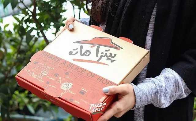پیتزا هات ستاری: جعبه پیتزا باز شده با لوگوی پیتزا هات و شعار "SHARE A SLICE OF HOPE" در دست مشتری. سفارش آنلاین پیتزا در تهران.