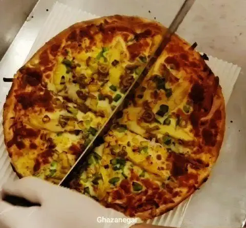 تصویر پیتزای تازه با خمیر طلایی و تاپینگ‌های ذرت و سبزیجات که با چاقو برش داده می‌شود، در پیتزا و کباب ترکی علی بابا.
