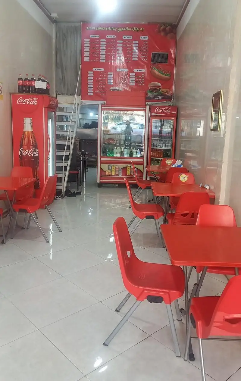 فضای داخلی پیتزا ساندویچی جوانان با میز و صندلی‌های قرمز، یخچال‌های نوشابه کوکاکولا، منوی غذاهای متنوع فست فود و پیتزا روی دیوار.