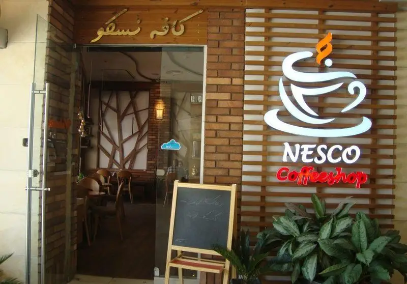 تصویر نمای بیرونی و ورودی کافه رستوران نسکو. تابلوی NESCO Coffeeshop، لوگوی فنجان قهوه، دیوار آجری و دکور چوبی مدرن.