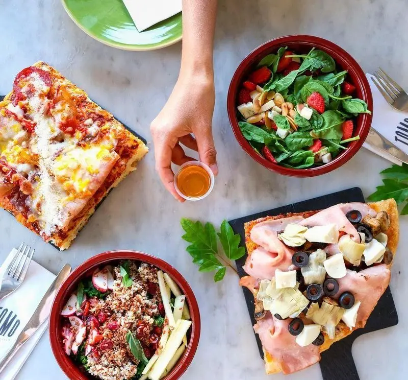 میز غذای فست فود تومو با دو پیتزای دلپذیر، دو سالاد تازه و دست در حال سرو سس. تنوع غذایی سالم در فست فود تومو.