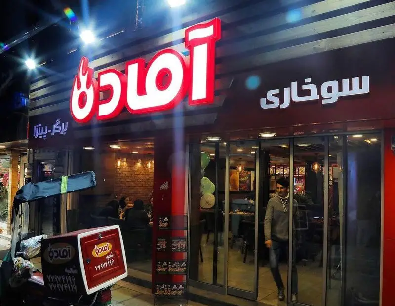 نمای شبانه و جذاب فست فود پیلوت (آماده) با تابلوی نئون روشن، ارائه دهنده انواع برگر، پیتزا و مرغ سوخاری به همراه خدمات تحویل.