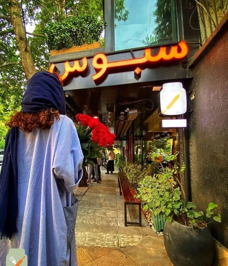 یک نفر با دسته گل رز قرمز در حال ورود به رستوران سبوس. تابلوی نورانی 'سبوس' و فضای سبز بیرونی، جلوه ای دلنشین به ورودی بخشیده.