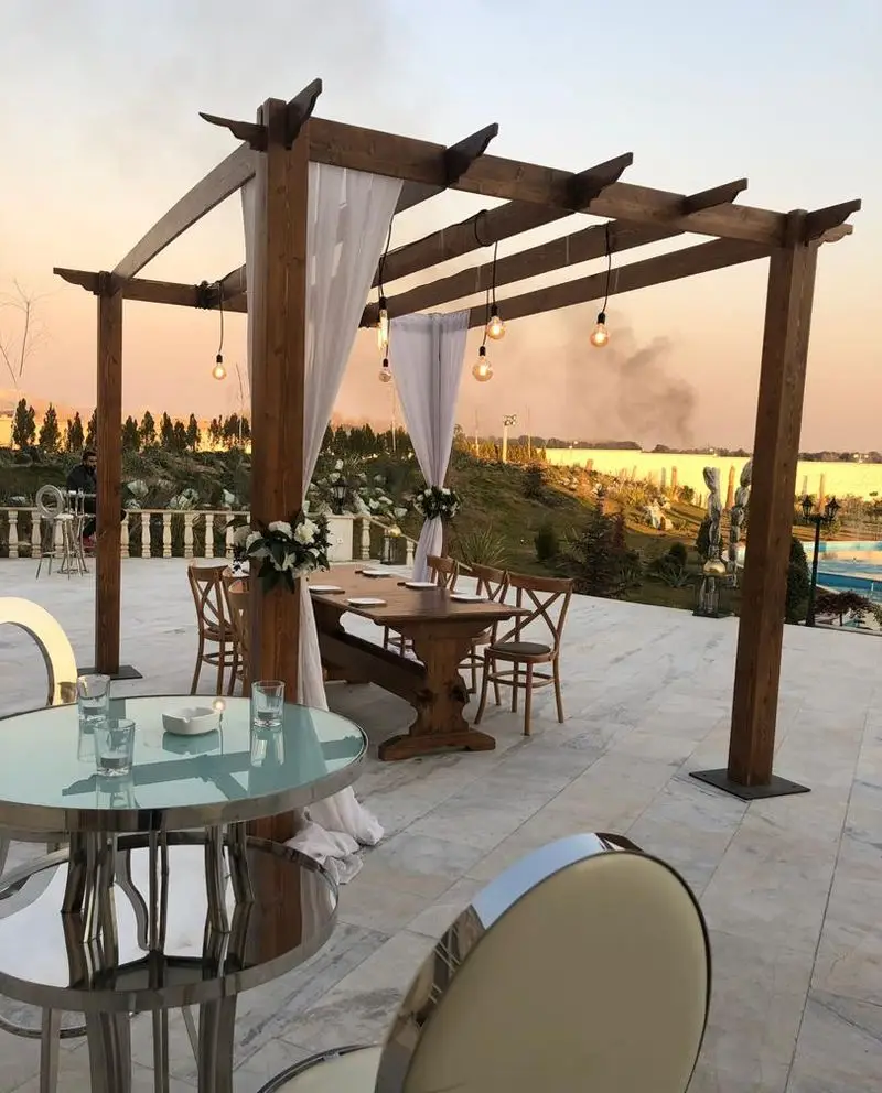 چیدمان رمانتیک میز و صندلی چوبی و دکوراسیون زیبا با پرگولای چوبی و نورپردازی، ایده‌آل برای اجاره ظروف و تشریفات بلادی در فضای باز.