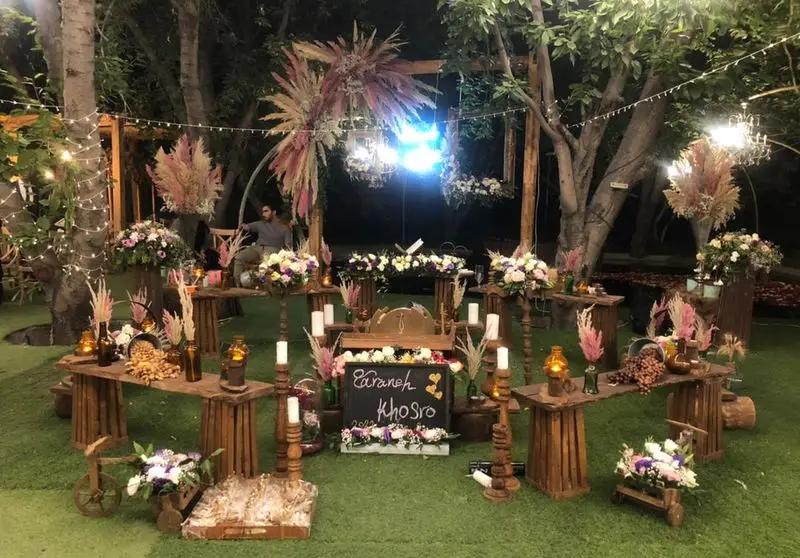چیدمان خاص و روستیک مراسم عقد و عروسی در فضای سبز باغ، شامل میزهای چوبی، گل‌آرایی و شمع‌آرایی با ظروف کرایه بلادی.