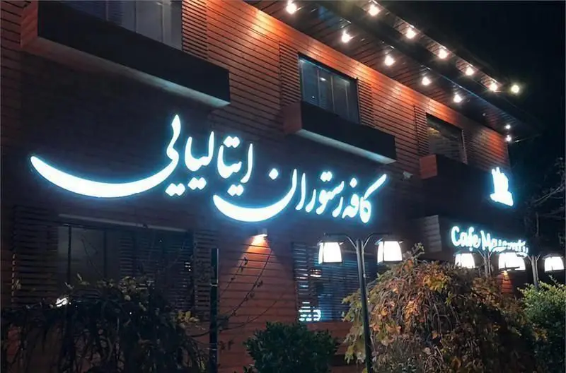 نمای شب کافه رستوران ایتالیایی مازراتی، یک کافی شاپ لوکس با تابلوی نئونی درخشان و طراحی چوبی مدرن و نورپردازی زیبا.
