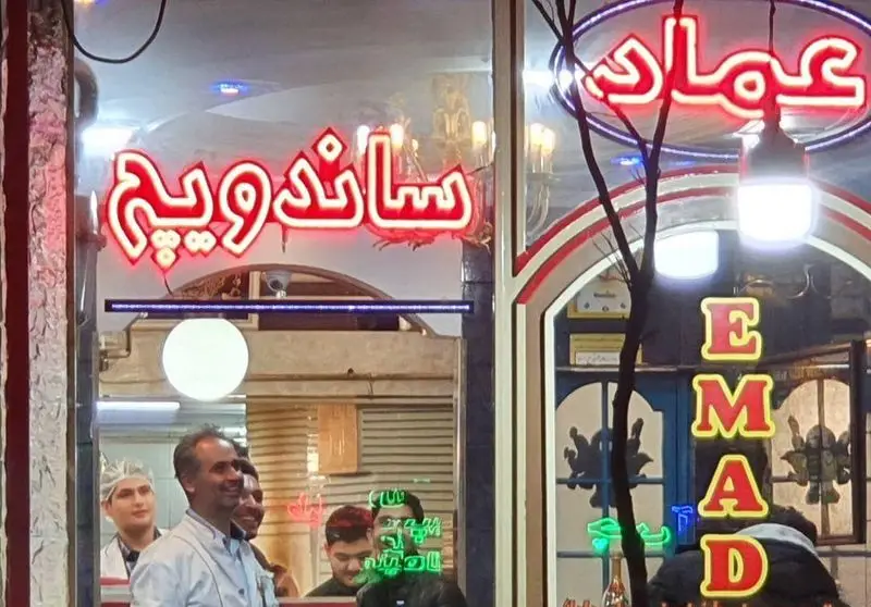 نمای شبانه و روشن مغازه فست فود پیتزا عماد با تابلوهای نئونی 'ساندویچ' و 'EMAD'. بهترین غذاهای فست فود را اینجا بیابید.