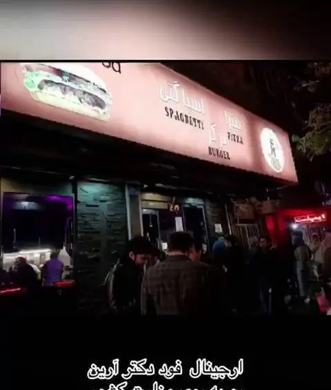 تصویر شبانه از نمای بیرونی فست فود "اورجینال فود دکتر آرین" با منوی متنوع شامل پیتزا، برگر و اسپاگتی.