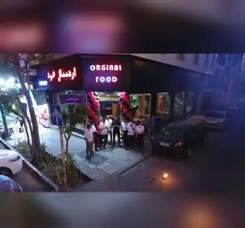 نمای شب از رستوران فست فود اورجینال (پیتزا دکتر آرین) با تابلوهای نئونی و کارکنان در حال استقبال مهمانان.