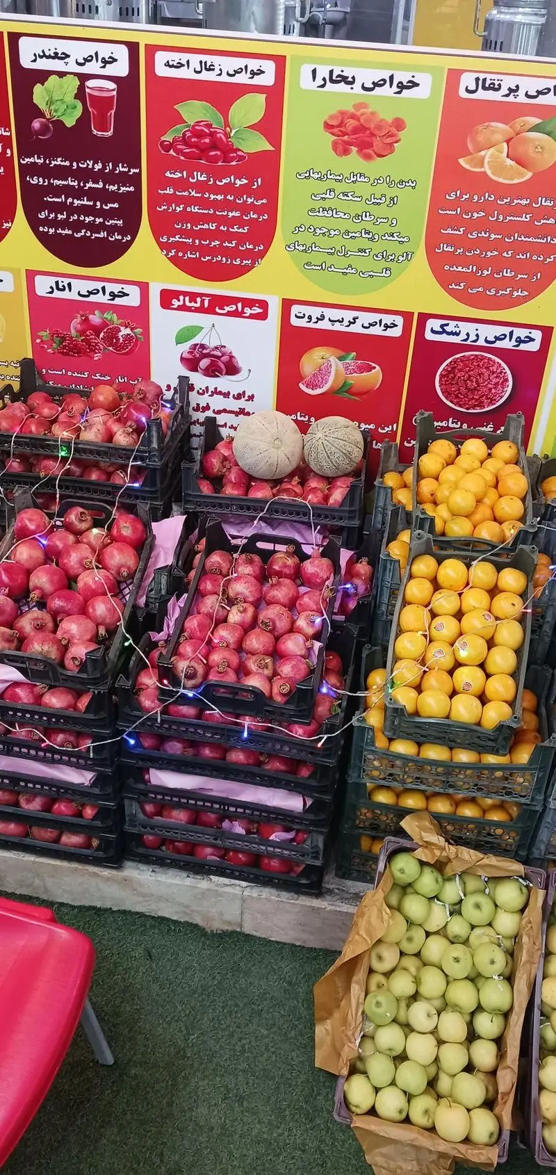 غرفه میوه ننه آقا در شهرری، نمایش میوه های تازه از جمله انار، پرتقال و سیب با تابلوهای خواص سلامتی میوه ها.