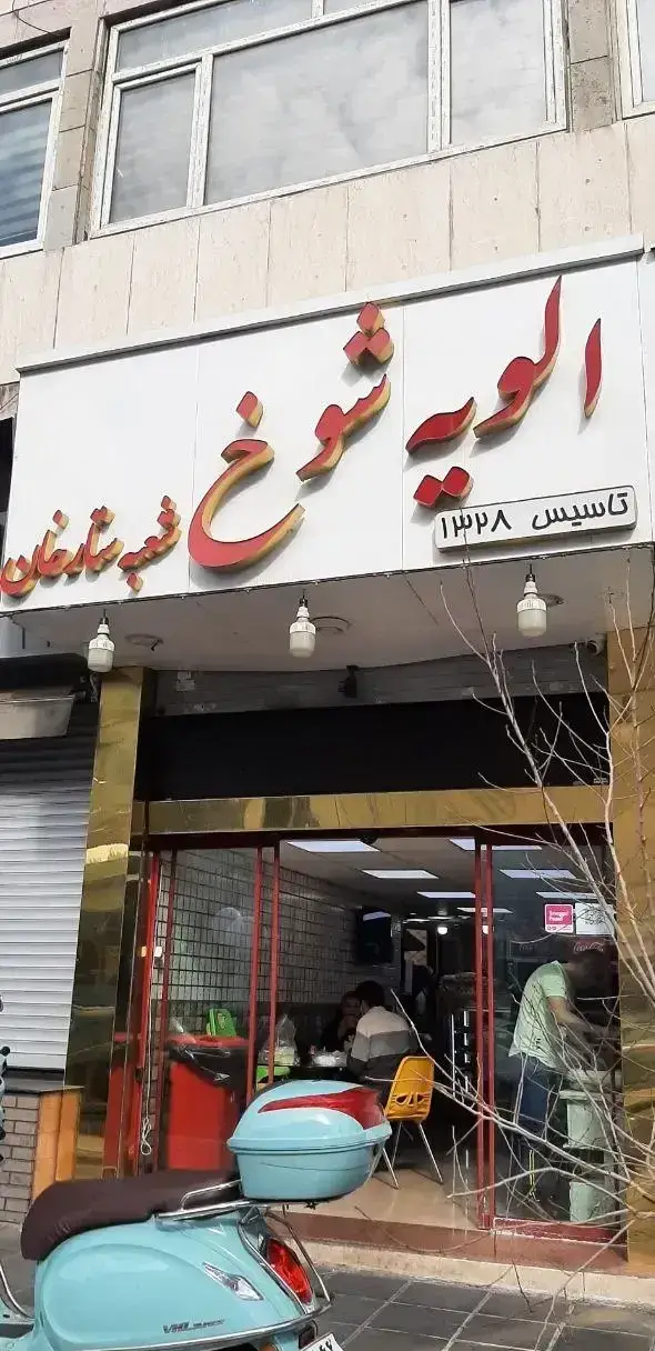 تصویر نمای بیرونی ساندویچی الویه شوخ شعبه ستارخان؛ فست فود معروف تهران با تابلوی بزرگ و اسکوتر آبی رنگ در جلوی مغازه