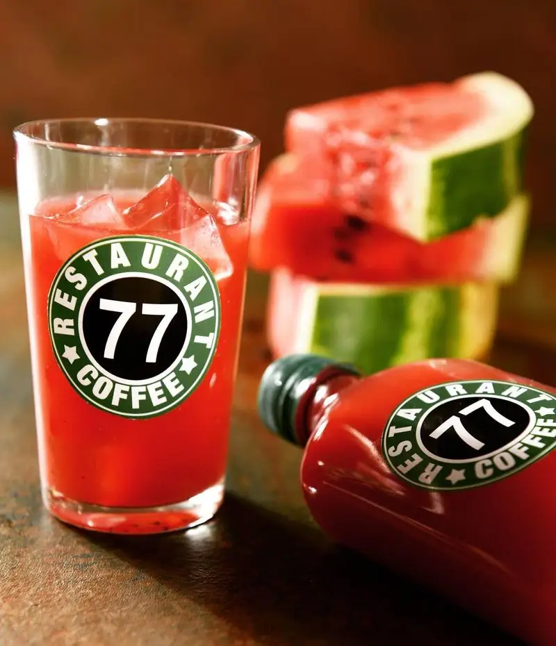 نوشیدنی آب هندوانه خنک در لیوان و بطری با لوگوی رستوران و کافه ۷۷، کنار برش‌های تازه هندوانه. ایده‌آل برای تابستان.