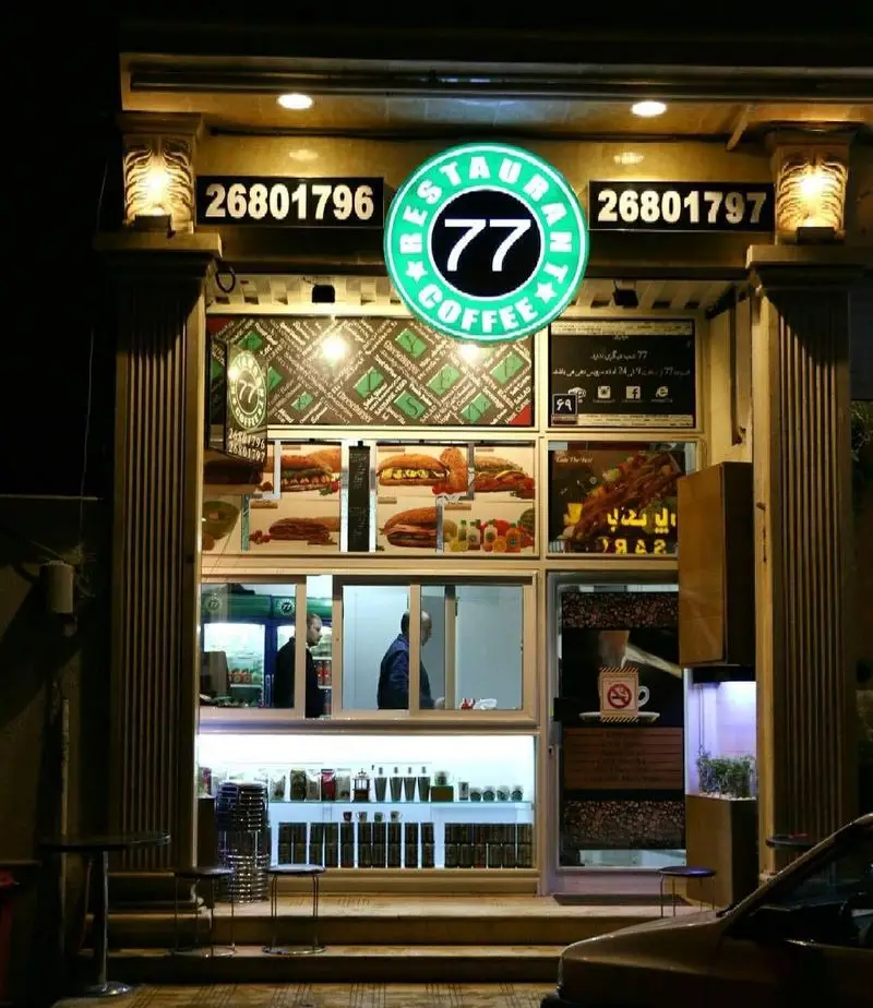 نمای شبانه رستوران و کافه ۷۷ با غذاهای فست فود و قهوه، با شماره تماس ۲۶۸۰۱۷۹۶ و ۲۶۸۰۱۷۹۷. محیط روشن و جذاب.